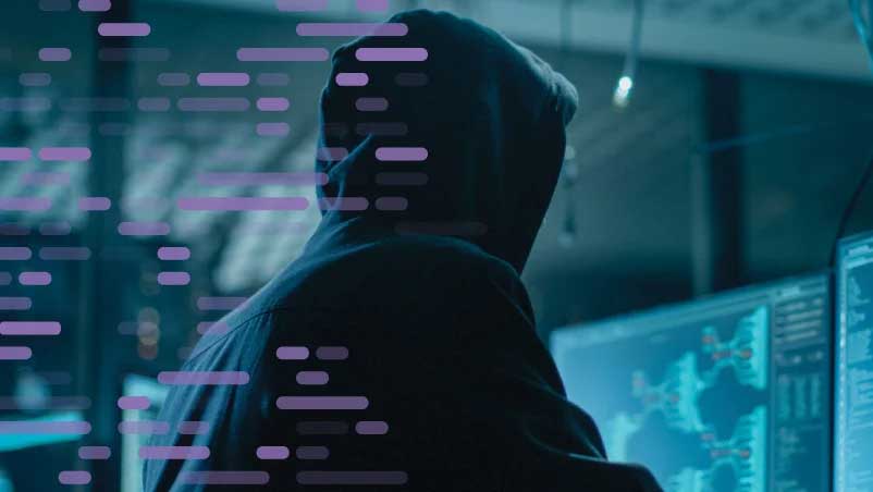 Hacker in a dark hoodie viewing glowing computer screens with data flowing.