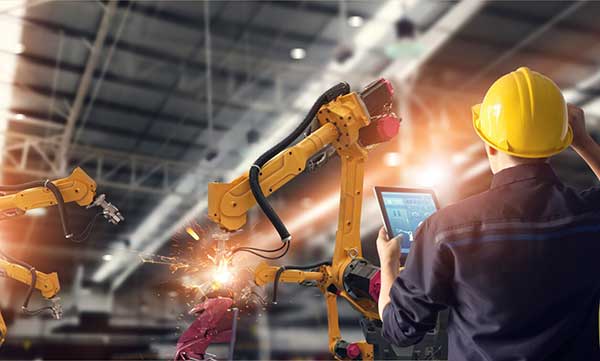 Engineer in a hard hat monitors yellow robotic arms welding in a bright factory.