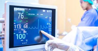Medical professional's gloved hand points to a monitor displaying patient vital signs.
