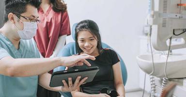 Doctor in mask showing a tablet to a smiling patient in a dental office.