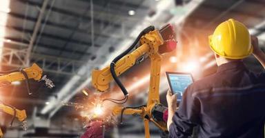 Engineer in a hard hat monitors yellow robotic arms welding in a bright factory.