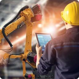 Engineer in hard hat controls a yellow robotic arm with a tablet in a factory.
