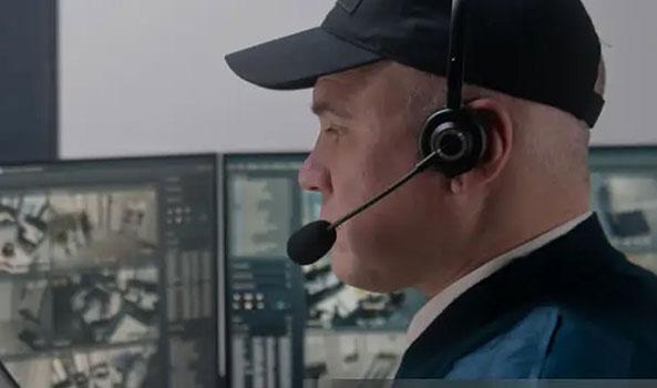 Man in black cap and headset monitoring security camera feeds on dual screens.