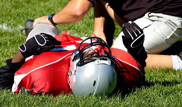 Injured football player in red jersey on field, trainer kneels beside him.