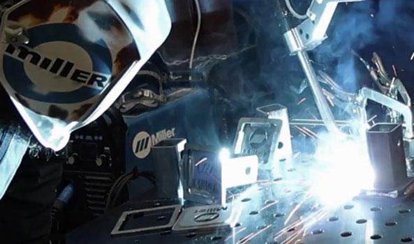 Welder in Miller helmet and gloves, sparks flying from active welding on metal parts.